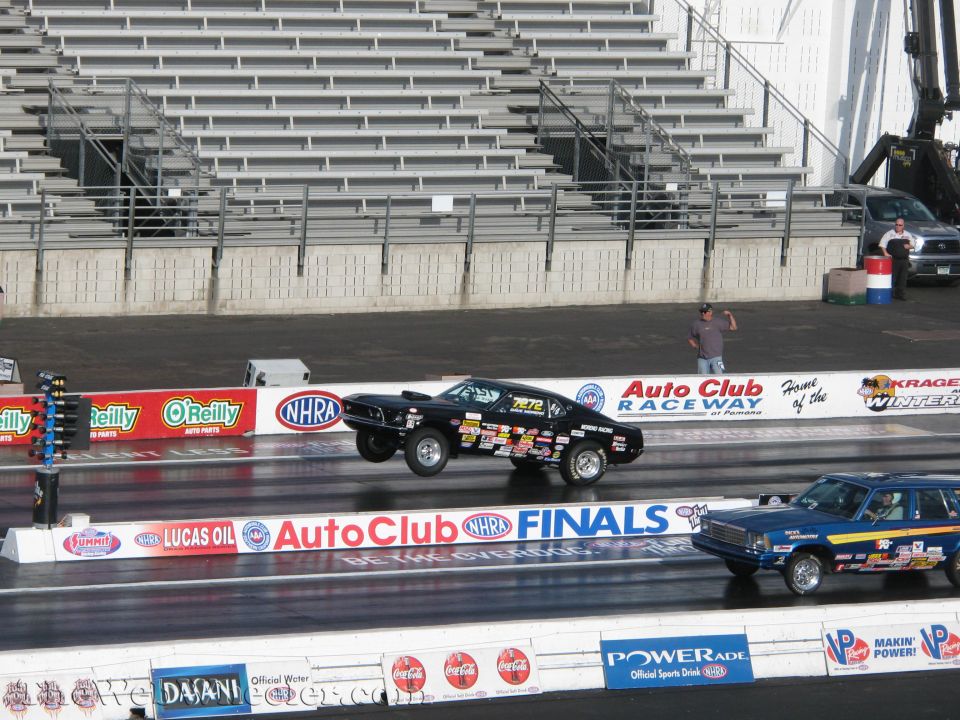 Black drag car doing a wheelie next to a blue station wagon on a drag strip
