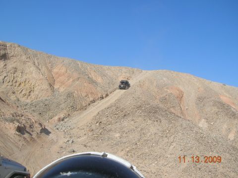 1985 Ford Bronco - A dark SUV navigates a challenging dirt incline in a desert landscape, with another vehicle in view.