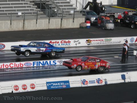 1968 Ford Mustang - Two drag cars compete on a drag strip, with the red car in the foreground and a blue car in the background.