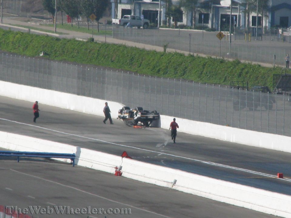 Overturned race car on track with emergency personnel nearby