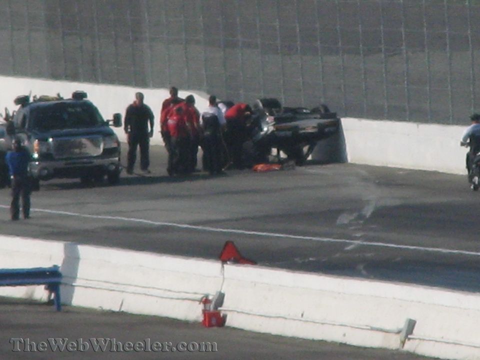 Overturned race car on track with safety crew and a GMC pickup truck nearby. A motorcycle is visible on the right