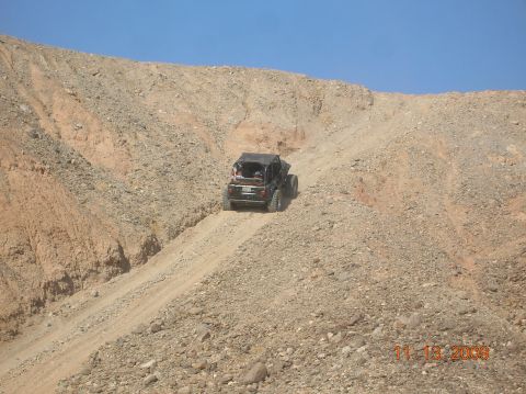 1985 Ford Bronco - A black off-road vehicle navigates a steep, rocky incline under a clear blue sky.