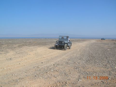1985 Ford Bronco - A gray Jeep CJ-5 navigates a dusty desert road under a clear blue sky.