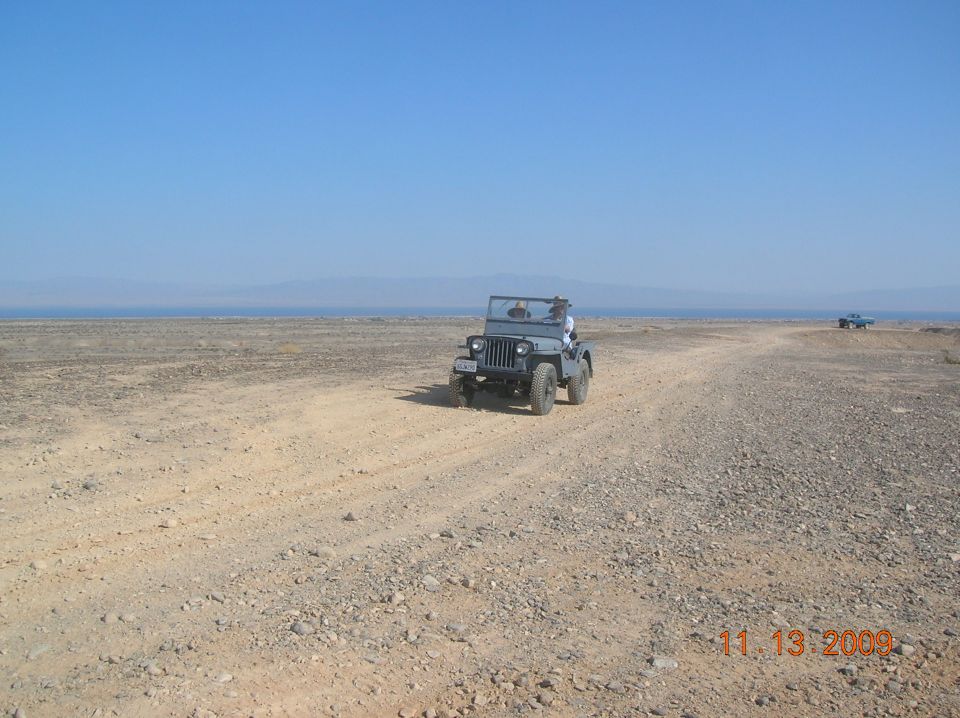 Gray Jeep CJ-5 driving on a dirt road with a blue truck in the distance