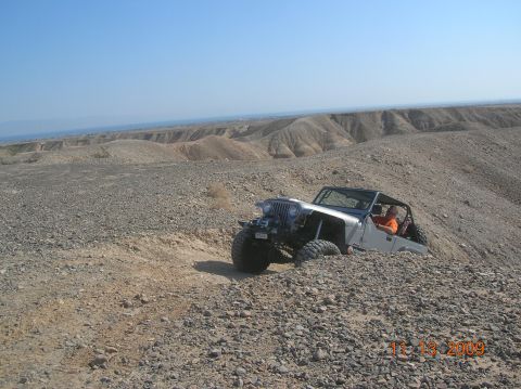 1985 Ford Bronco - A silver Jeep Wrangler tackles a challenging off-road trail in a barren, rocky desert environment.