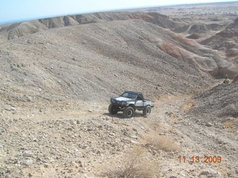 1985 Ford Bronco - A modified pickup truck navigates a desert landscape with hills and dry brush.