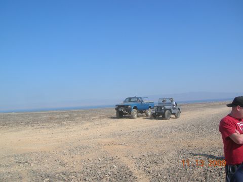 1985 Ford Bronco - Two off-road vehicles are parked on a dusty, rocky terrain under a clear blue sky.