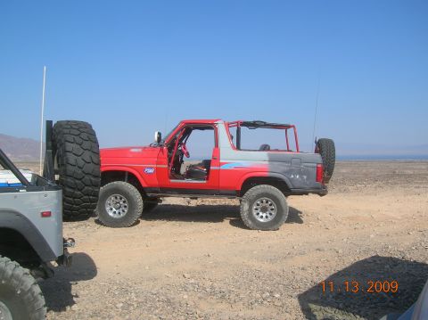 1985 Ford Bronco - A red and grey 1985 Ford Bronco is parked in a desert landscape with another vehicle partially visible.