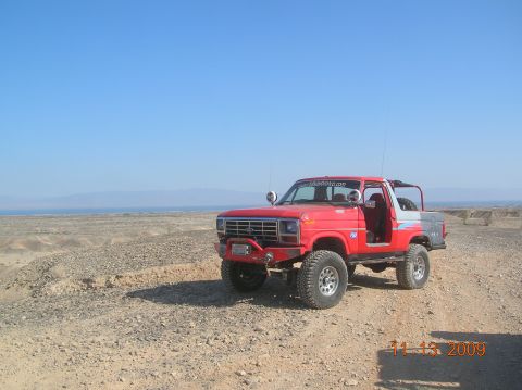 1985 Ford Bronco - Modified 1980s Ford Bronco sits on a dirt road under a clear blue sky with a desert landscape behind it.