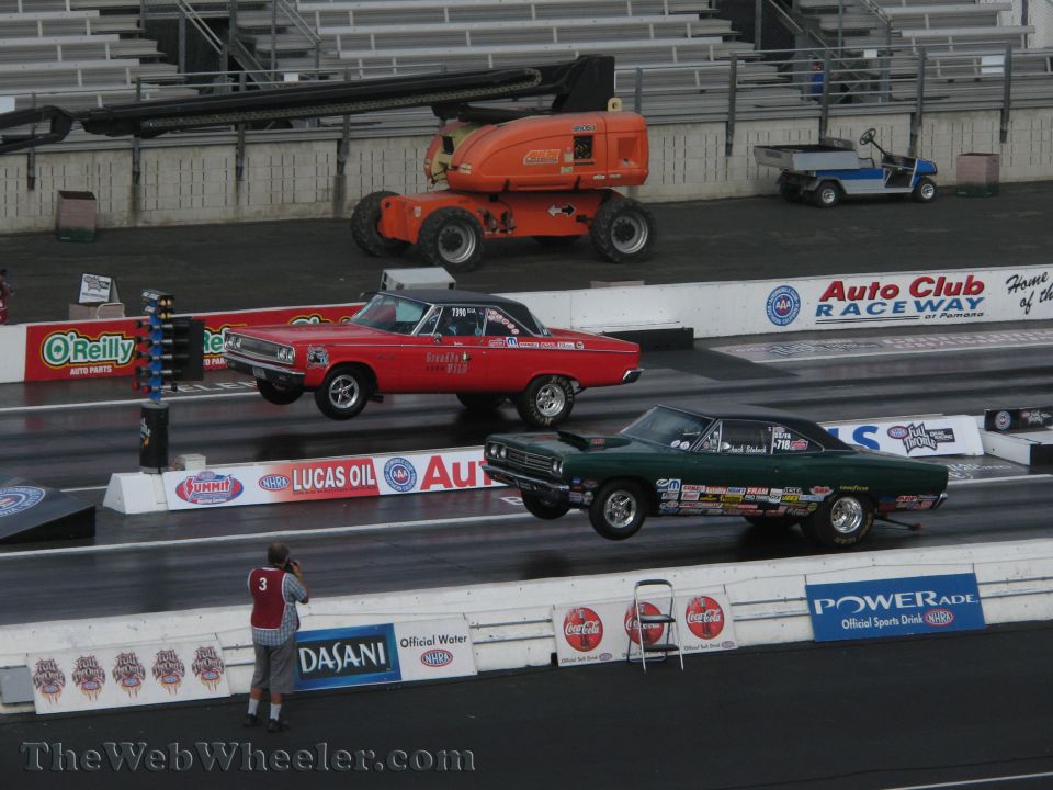 two muscle cars drag racing, one red, one green, both lifting front wheels off the ground