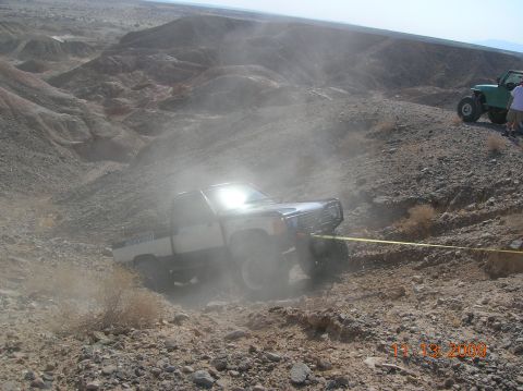 1985 Ford Bronco - Two off-road vehicles navigate a steep, dusty trail in a desert landscape.