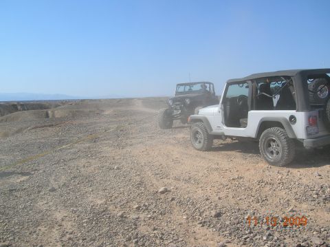 1985 Ford Bronco - Two Jeeps drive on a dusty, rocky path in a desert landscape.