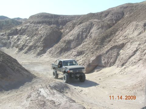 1985 Ford Bronco - Pickup truck navigates a dusty desert trail with steep, rocky canyon walls on either side.