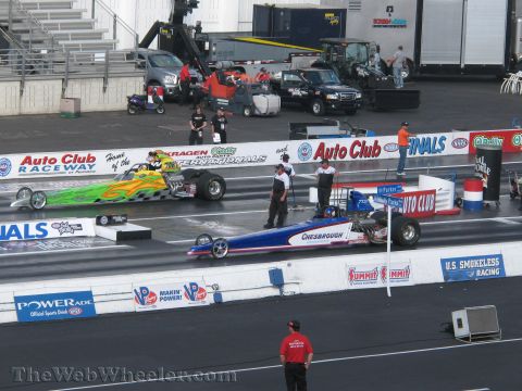 1968 Ford Mustang - Two dragsters are lined up on a race track, with various support vehicles and grandstands visible in the background.