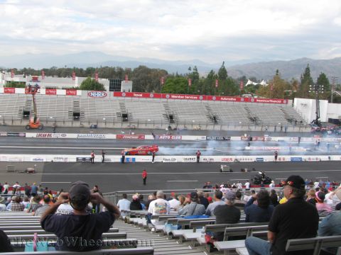 1968 Ford Mustang - A drag race is underway at a stadium with spectators in the stands, mountains visible in the distance.