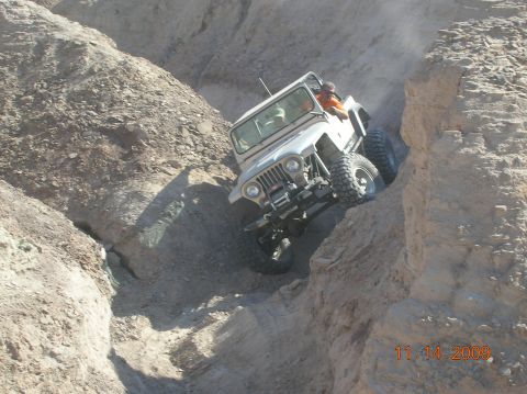 1985 Ford Bronco - A Jeep CJ-7 tackles a challenging off-road trail in a narrow canyon, kicking up dust as it climbs.