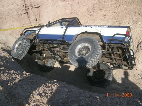 1985 Ford Bronco - A modified off-road truck navigates a steep dirt embankment with its wheels at extreme angles.