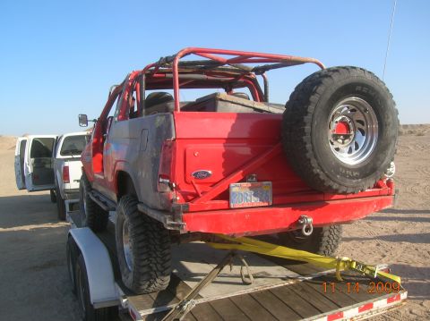 1985 Ford Bronco - A red off-road SUV is loaded onto a trailer, with a white SUV following behind it in a desert landscape.