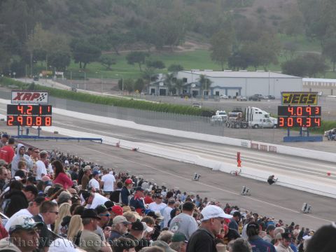 1968 Ford Mustang - A drag strip with spectators in the stands and electronic scoreboards displaying race times.