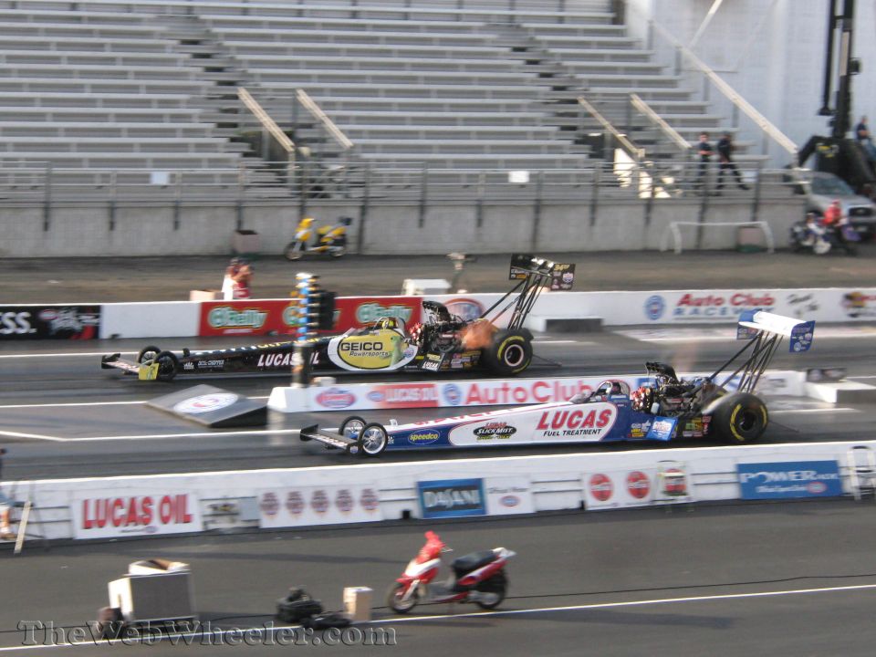 Two drag racing cars compete side-by-side, one with flames erupting from its engine