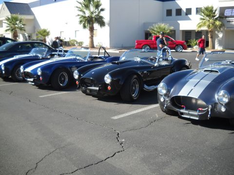 1986 Ford Bronco II - Multiple Shelby Cobra-style sports cars are parked in a lot, with a red pickup truck visible behind them.