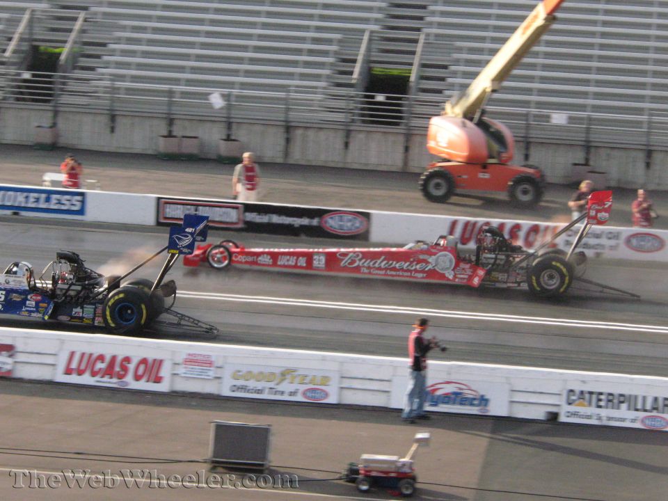 Two drag racing cars on a track, one red with Budweiser sponsorship, the other blue and black, with a cherry picker in the background