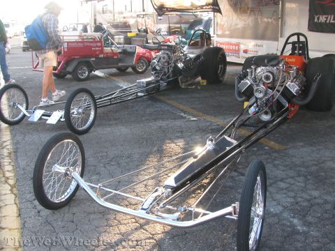 1968 Ford Mustang - Two dragsters are parked on asphalt, with a person and golf carts in the background.