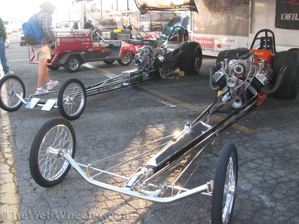 Two dragsters with exposed engines and chrome frames on asphalt