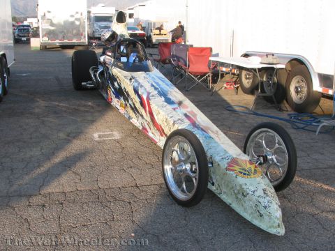 1968 Ford Mustang - A dragster with a red, white, and blue paint scheme is parked on an asphalt surface with trailers and chairs in the background.