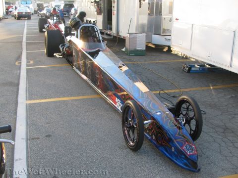 1968 Ford Mustang - A dragster with custom flame graphics is parked in a pit area next to trailers and other vehicles.