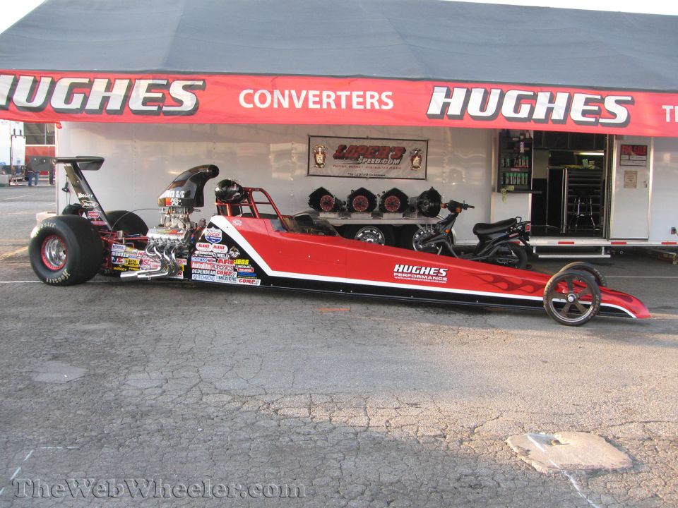 Red dragster with flames and sponsor decals, parked in front of a Hughes Converters trailer