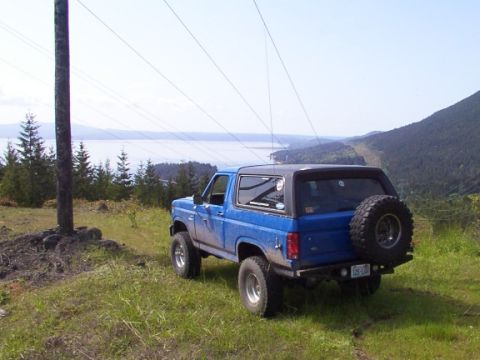 1980 Ford Bronco - A blue 1980 Ford Bronco is parked on a grassy overlook with a scenic view of a lake and mountains.