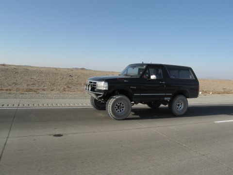 1986 Ford Bronco - A black Ford Bronco is seen driving on a highway under a clear blue sky with a desert landscape.