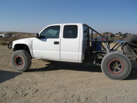 1986 Ford Bronco - Modified white pickup truck with off-road tires and roll cage sits on a dirt surface.