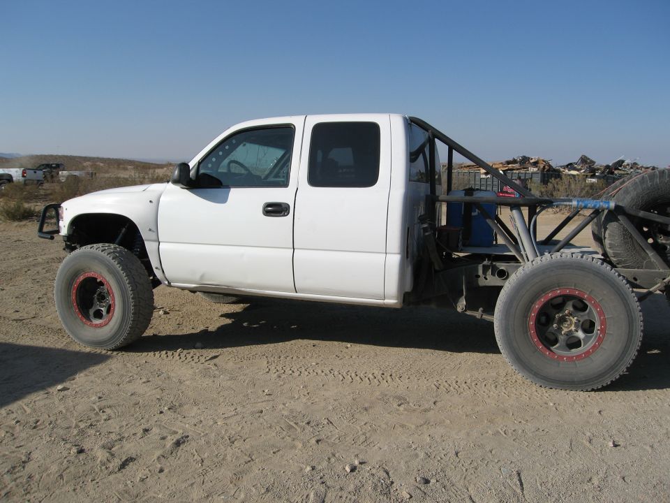 White pickup truck modified for off-road use, showing a roll cage and large tires