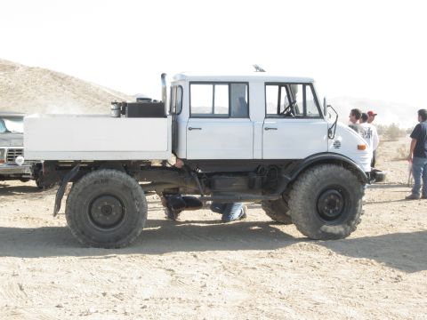 1986 Ford Bronco - A white crew cab flatbed truck with large off-road tires is parked on a sandy surface, with other vehicles and people in the background.