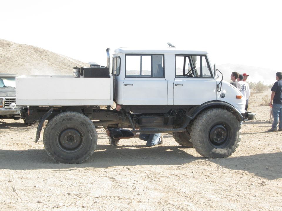 White flatbed truck with a crew cab, large tires, and a flatbed cargo area, parked on a dirt surface