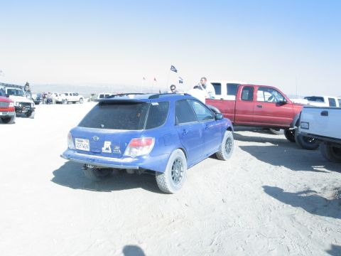 1986 Ford Bronco - A blue Subaru Outback wagon with aftermarket wheels and raised suspension is seen in a sandy environment alongside other vehicles.