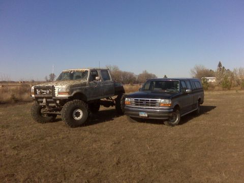 1994 Ford F-150 - Two Ford trucks, a heavily modified pickup and a Bronco, sit in a grassy field under a clear blue sky.