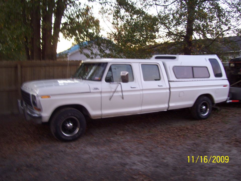 White Ford crew cab pickup with matching camper shell parked on gravel by a wood fence.
