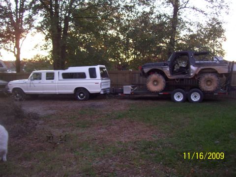 1978 Ford Bronco - Driver-side view of a white 1970s Ford crew cab pickup with a matching white bed cap towing a tandem-axle flatbed trailer loaded with a mud…
