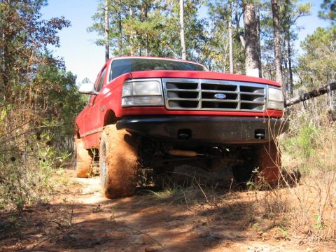 1995 Ford F-150 - This red Ford pickup truck is driving off-road through a wooded area, with mud caked on its tires.