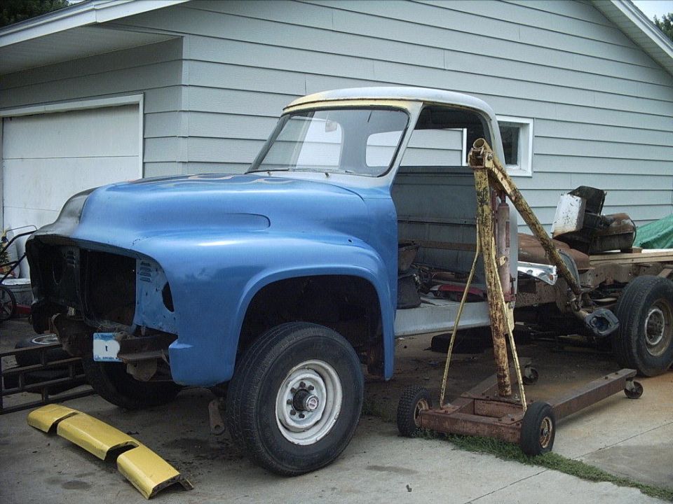 Blue 1954 Ford F-100 under restoration, missing grille and bed, with engine hoist beside a gray garage