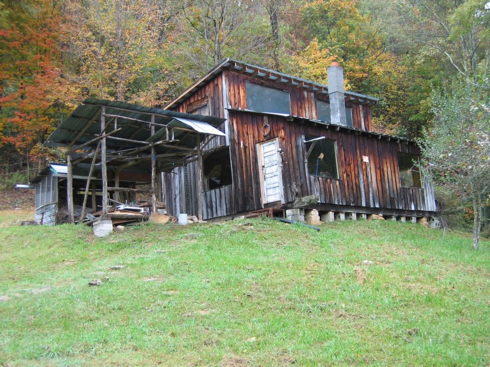 Dilapidated wooden building with a corrugated metal roof extension, set on a grassy slope with autumn trees in the background