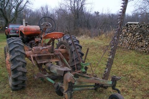 2000 Chevrolet Tahoe - An old orange tractor with a sickle bar mower is parked in a grassy area next to a woodpile, with a red pickup truck in the distance.