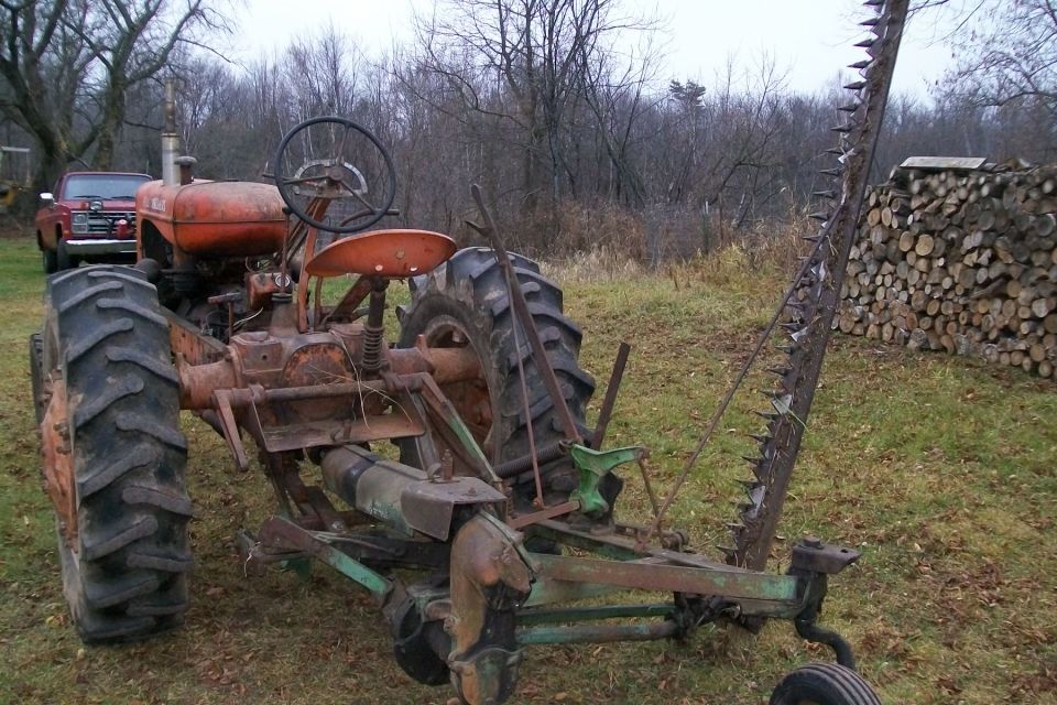 Orange tractor with a sickle bar mower attachment, viewed from the front-left. A red pickup truck is visible in the background