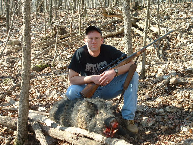 Man in black cap kneeling over a dead boar in rocky woods with a scoped rifle