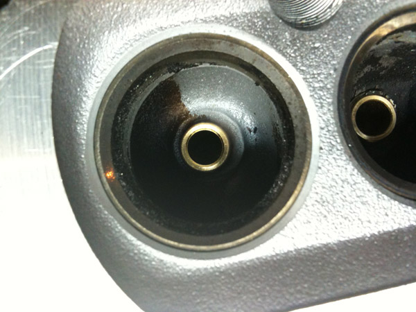 Close-up of an aluminum cylinder head combustion chamber with valve removed, showing brass valve guide