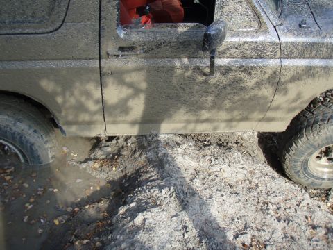 1985 Ford Bronco - Mud-covered 1985 Ford Bronco is stuck in a muddy ditch, with its front tire submerged.