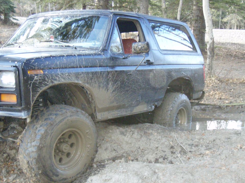 Mud-splattered dark blue 1980s Ford Bronco stuck in mud, front passenger side wheel deep in muck